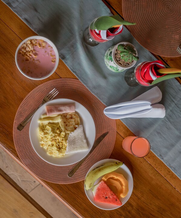 Un desayuno con un plato de huevos, tostadas y una porción de pastel, acompañado de un bol de fruta y un vaso de batido rosa.