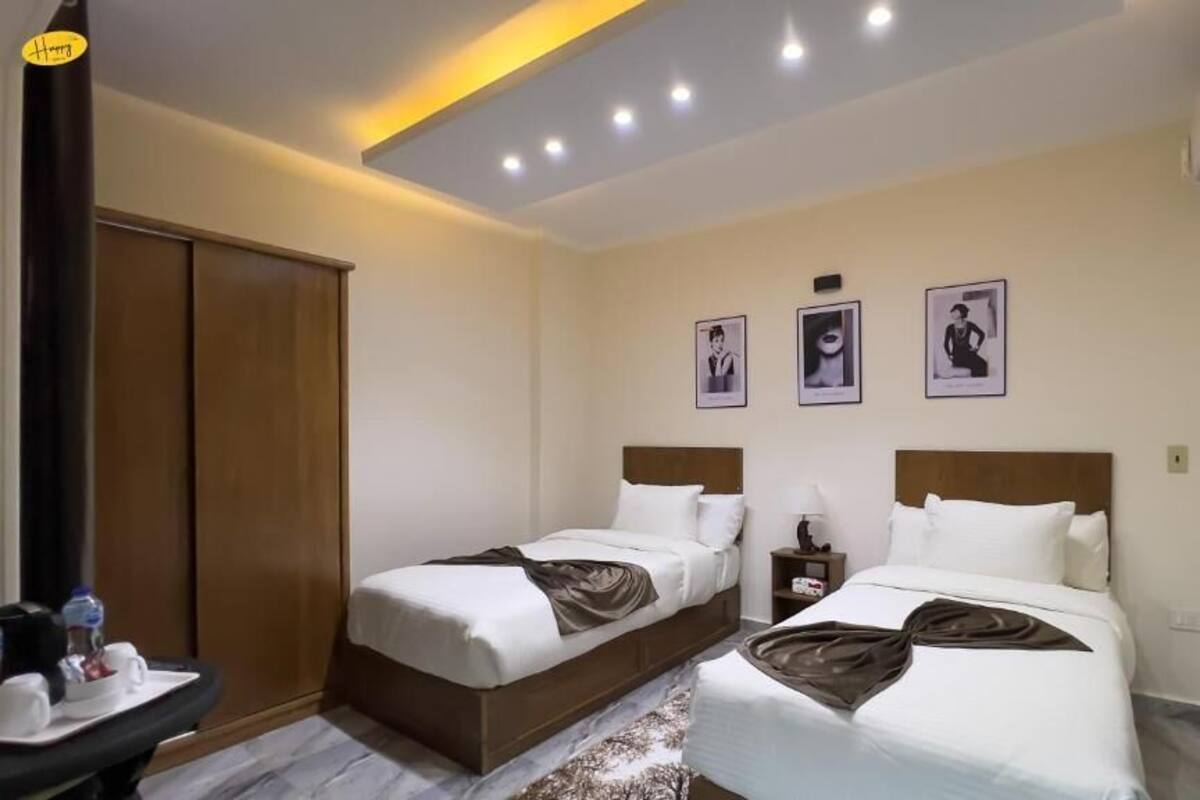 A hotel room with two beds, a wardrobe, a bedside table with a lamp, and framed pictures on the wall.