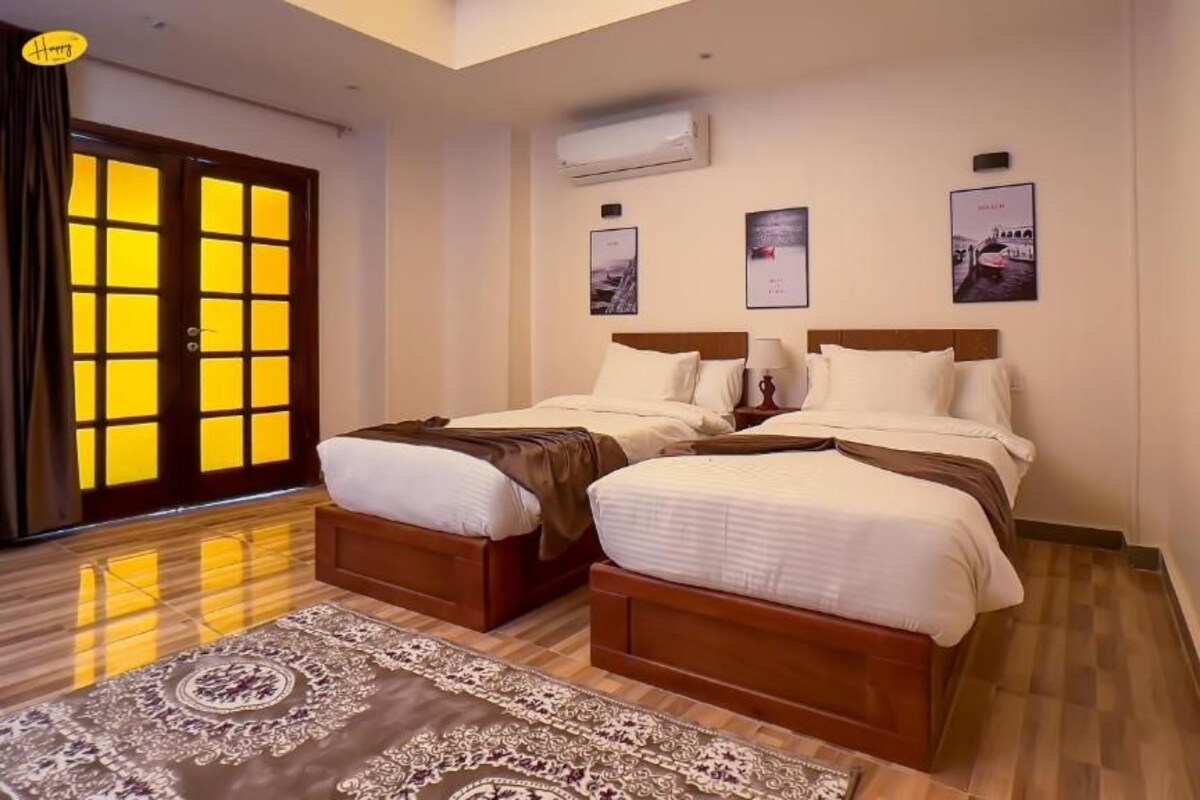 Habitación de hotel con dos camas, suelo de madera, un ventanal grande con cortinas y aire acondicionado instalado en la pared.