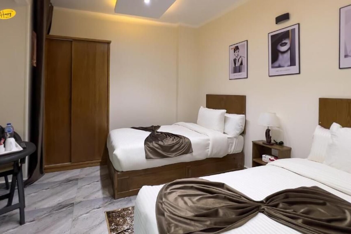 A hotel room with two beds, a wooden wardrobe, a desk with a chair, and a nightstand with a lamp.