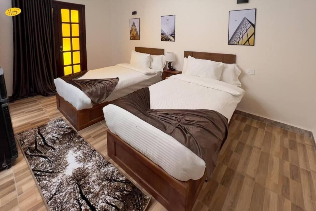 A hotel room with two beds, a wooden floor, a window with curtains, and three framed pictures on the wall.