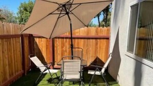 Un jardin avec une pelouse artificielle, une clôture en bois et un espace patio comprenant deux chaises et une table placés sous un grand parasol.