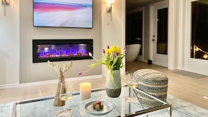 Una sala de estar moderna con un televisor de pantalla plana, una chimenea, una mesa de centro de vidrio con aperitivos y un rincón de descanso acogedor.