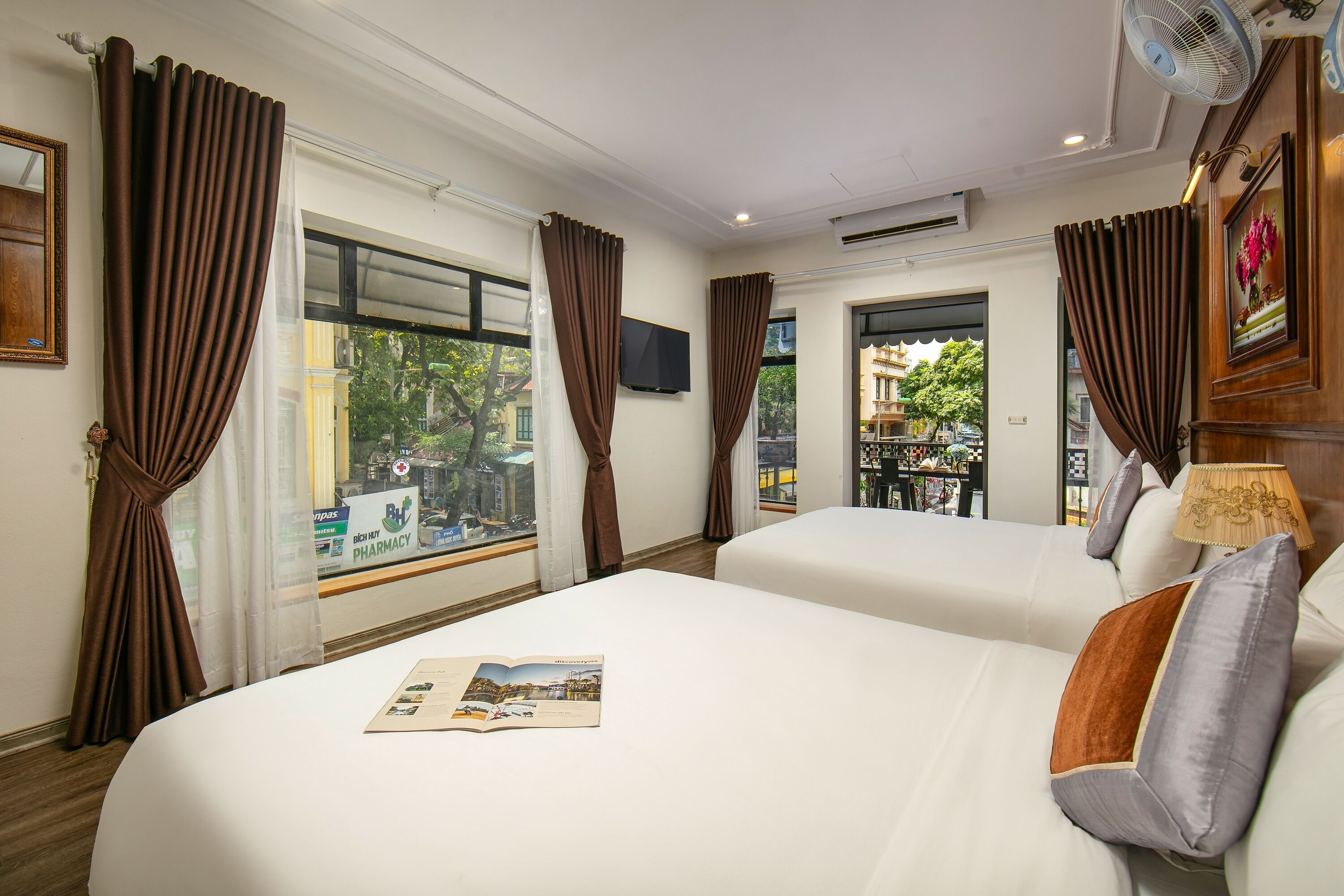 Habitación de hotel con dos camas, cortinas marrones, televisor montado y vista al exterior.