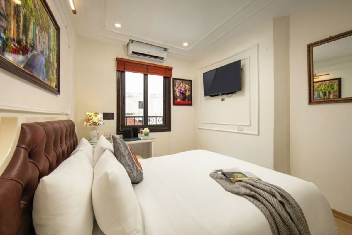 A hotel room with a large bed, a TV mounted on the wall, a window with a view, and a painting on the wall.