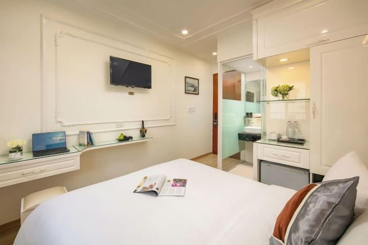 A hotel room with a bed, a desk with a laptop, a TV mounted on the wall, and a bathroom visible through a glass door.