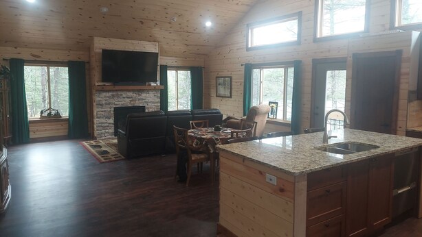 A log cabin interior with a stone fireplace, a television mounted above it, a dining area with a table and chairs, and a kitchen with a granite countertop and sink.