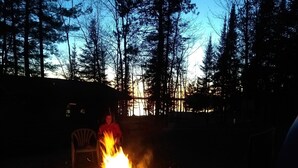 A campfire with logs burning in a metal container, surrounded by trees and a chair in a forested area.