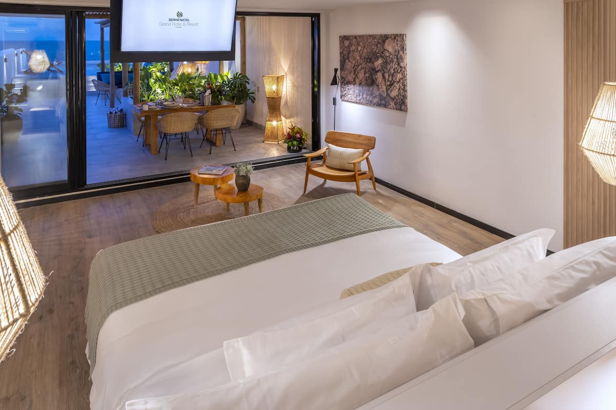 A hotel room with a bed, a dining area with chairs and a table, a view of the sea, and a large painting on the wall.