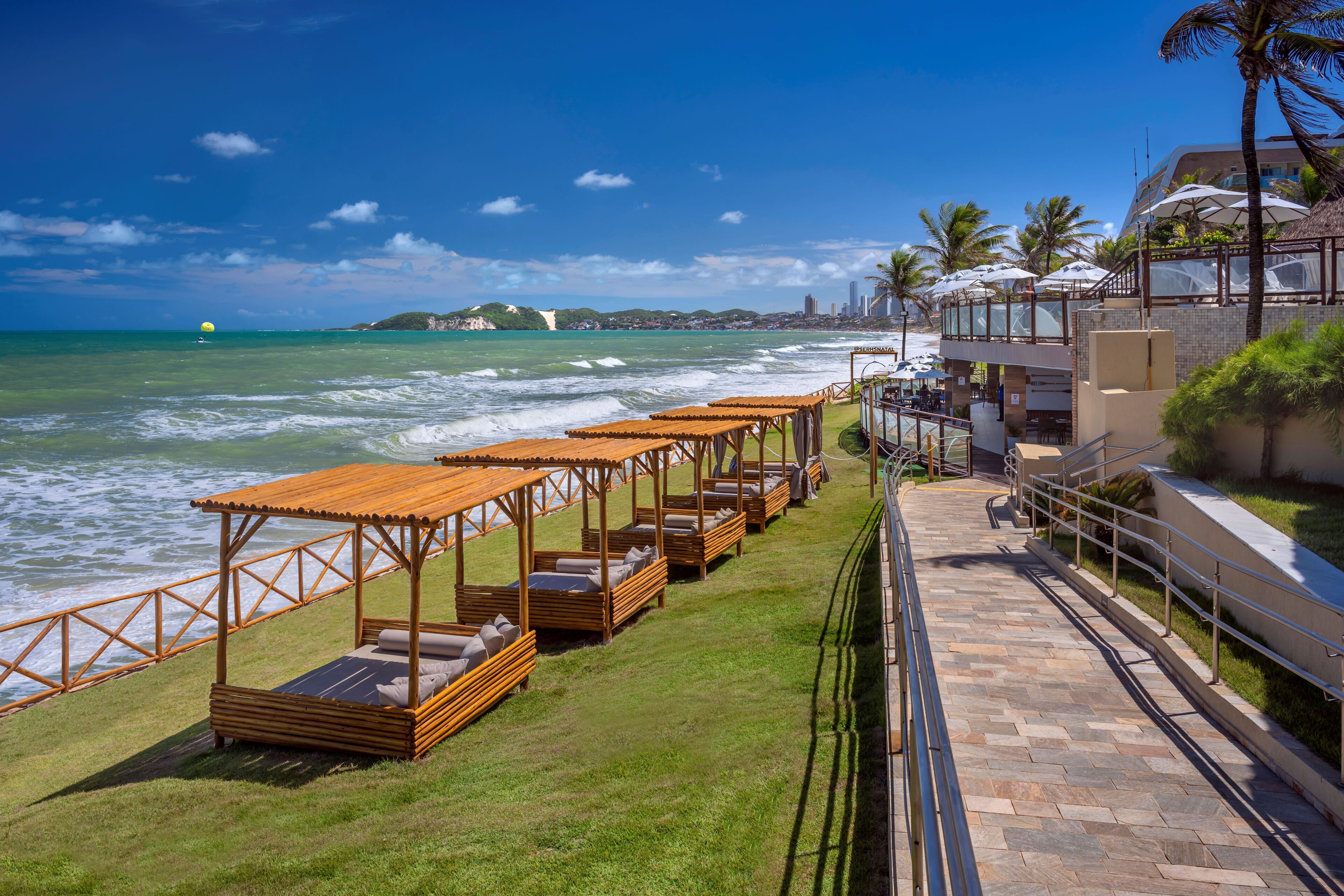Un hotel frente a la playa con zonas de estar al aire libre, un camino que lleva a la playa y vistas al océano y al horizonte urbano.