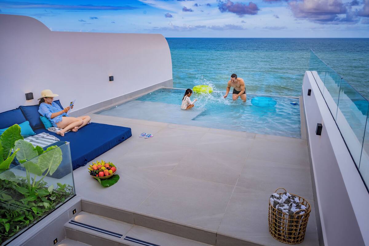 A modern infinity pool with a clear view of the ocean, a lounge area with a sofa and a small table, and a wicker basket with towels.