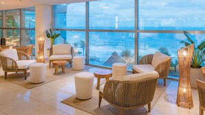 A modern lounge area with wicker furniture, white cushions, and large windows offering a view of the sea. - SERHS Experience Suites (Natal)