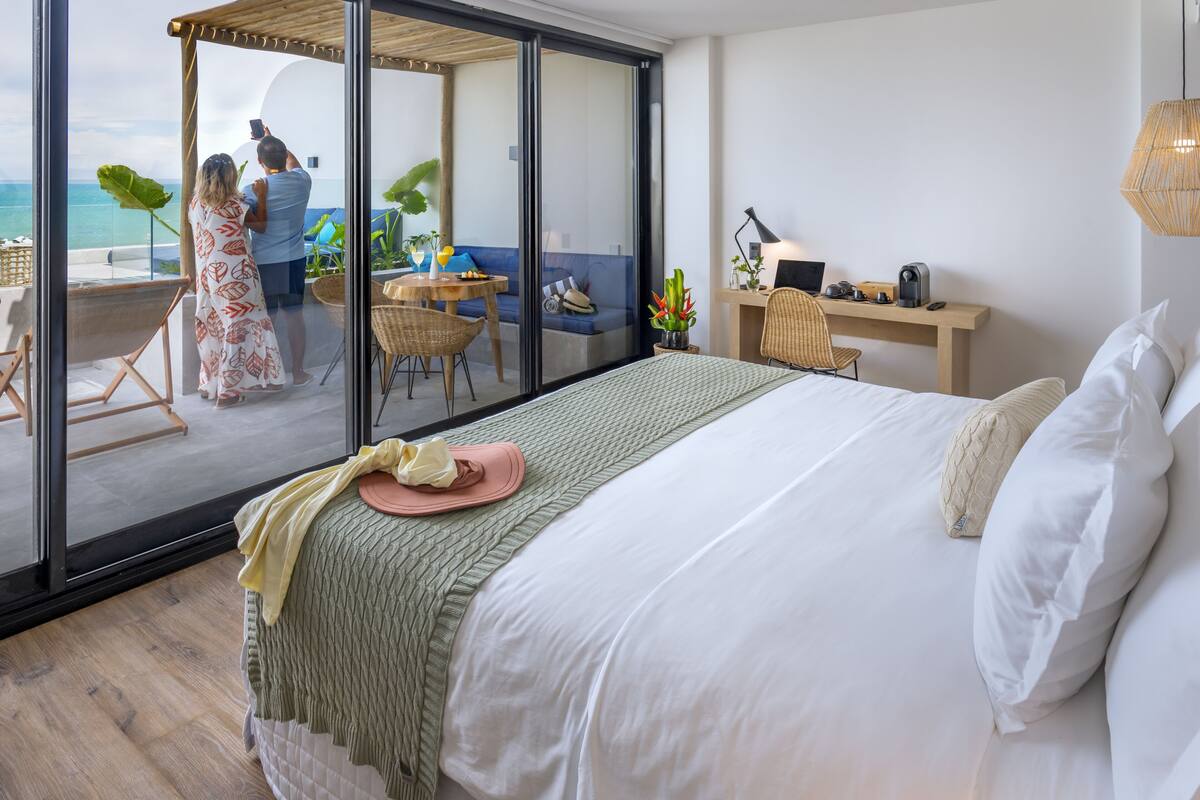A modern hotel room with a large bed, a desk, a chair, and a balcony with a view of the ocean.