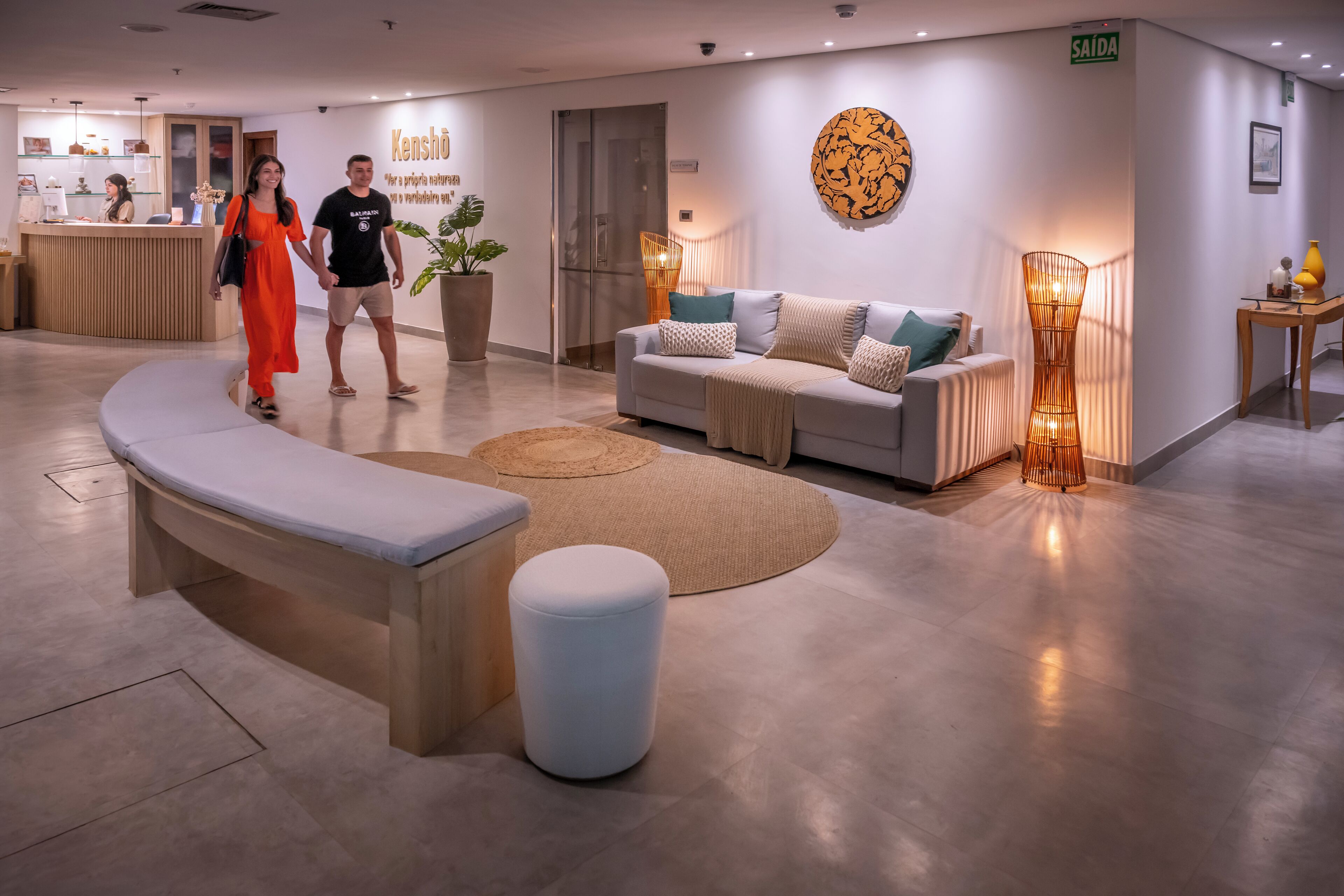 Un moderno vestíbulo de hotel con mostrador de recepción, un sofá con cojines, un tapete redondo y una lámpara de pie alta.