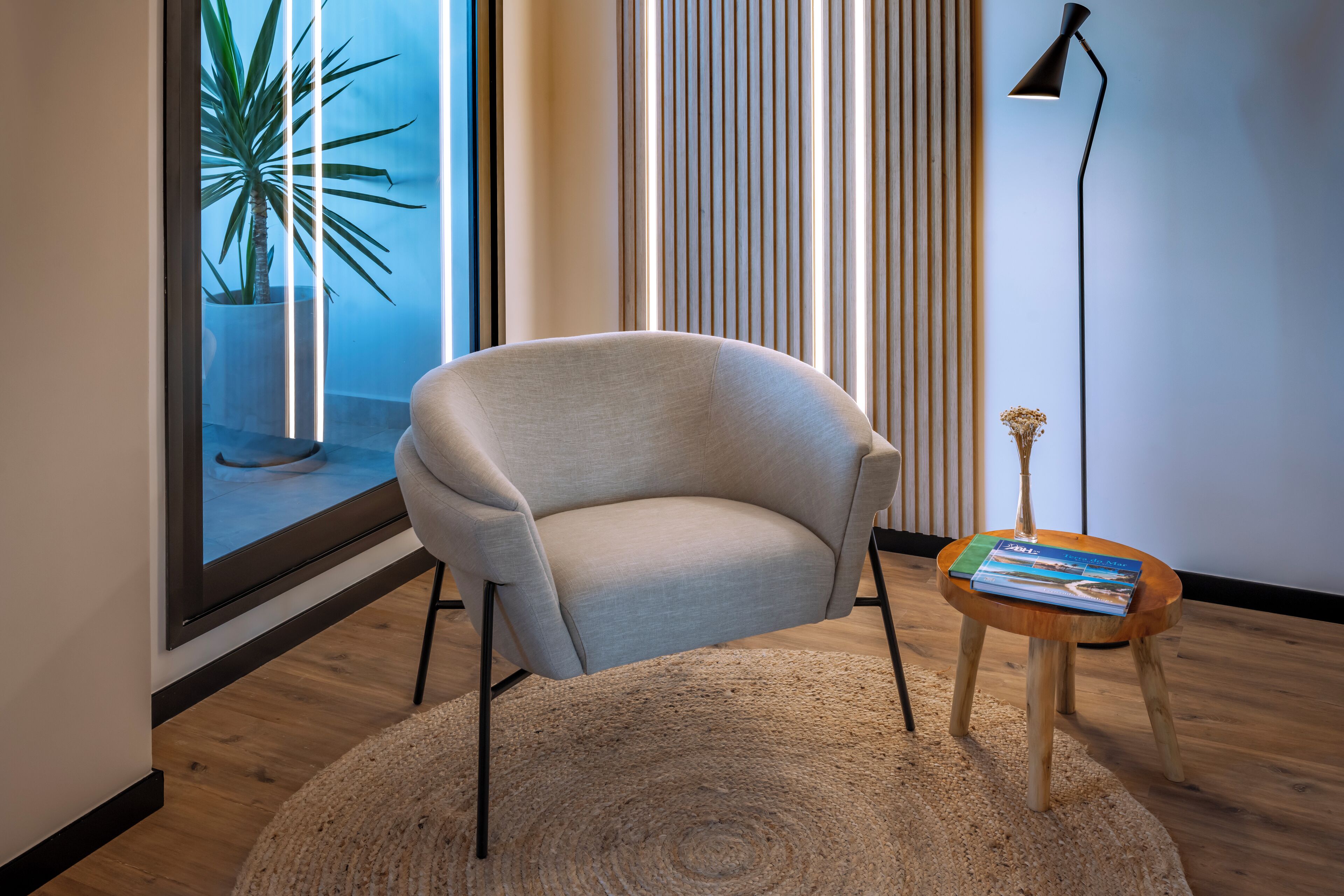 Una habitación moderna con un ventanal grande, un sillón cómodo, una mesita de madera con un libro y una lámpara de pie alta.
