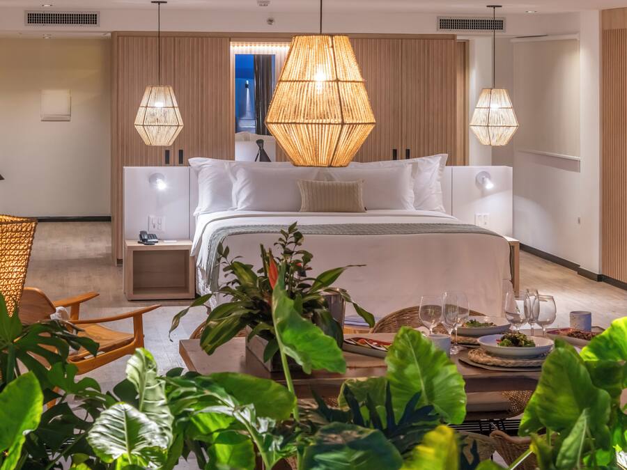 A modern hotel room with a large bed, two bedside tables, a dining area with a table set for two, and a large window.