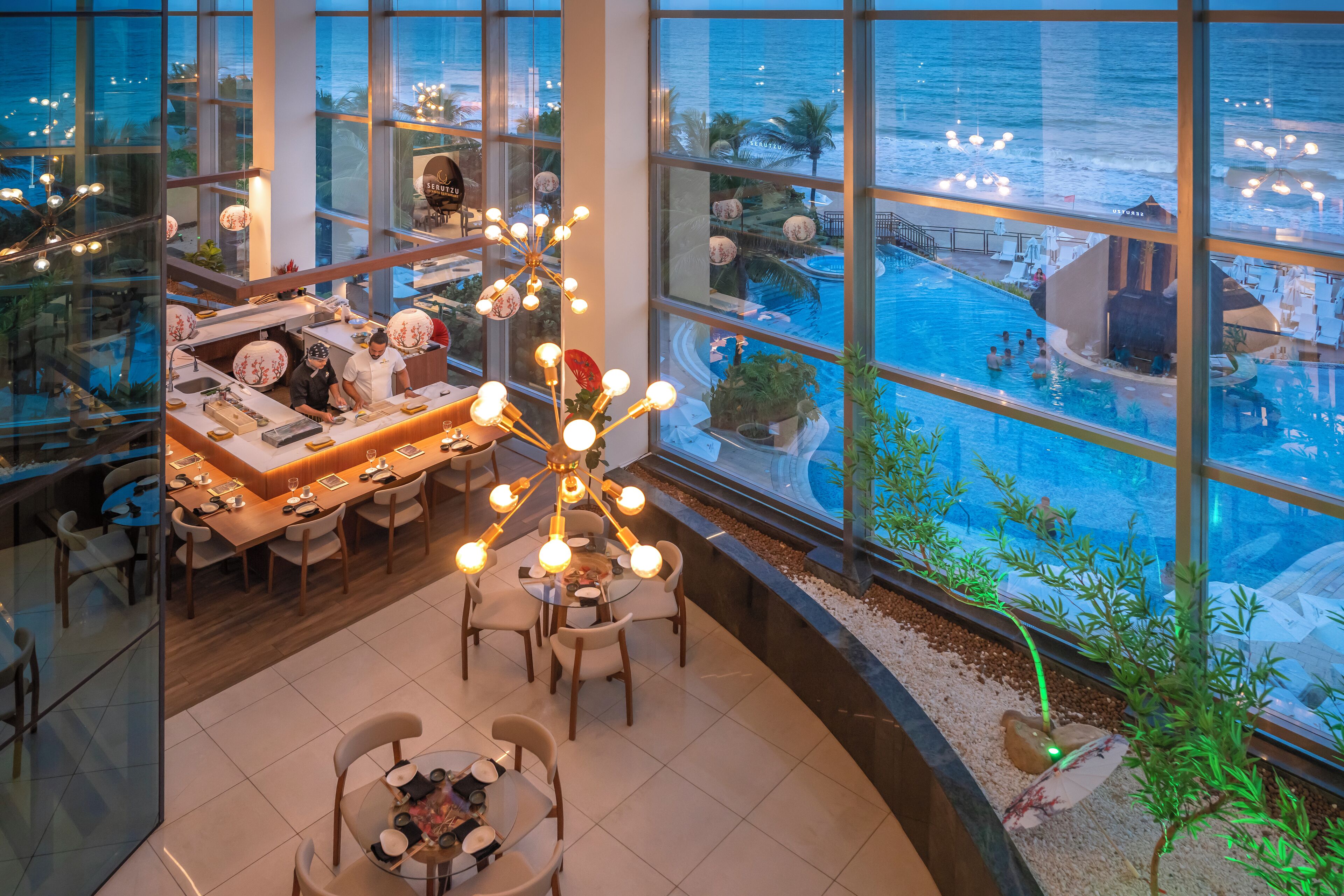 Un moderno vestíbulo de hotel con vista a una piscina, grandes ventanales y un área central de comedor.