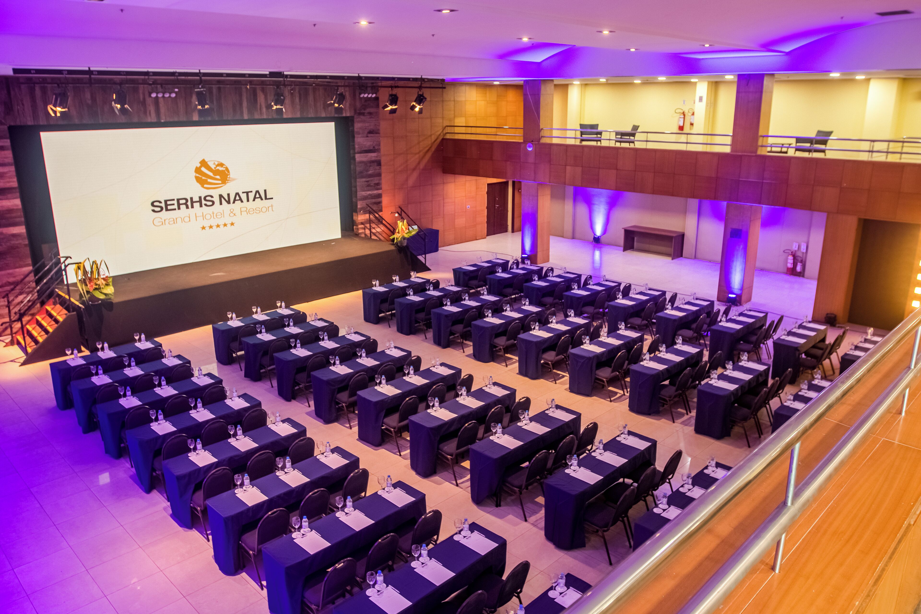 Un salón de conferencias con varias mesas y sillas, un escenario con una pantalla de presentación y una escalera que conduce a un nivel superior.