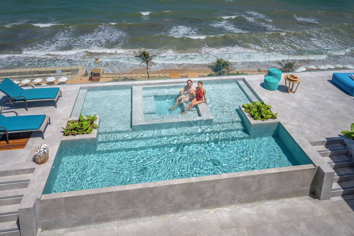 A rooftop pool with a view of the ocean, lounge chairs, and a small table with a drink.