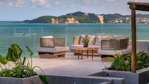 A terrace with wicker furniture, a table, and potted plants, overlooking a coastal view with mountains in the background. - SERHS Experience Suites (Natal)