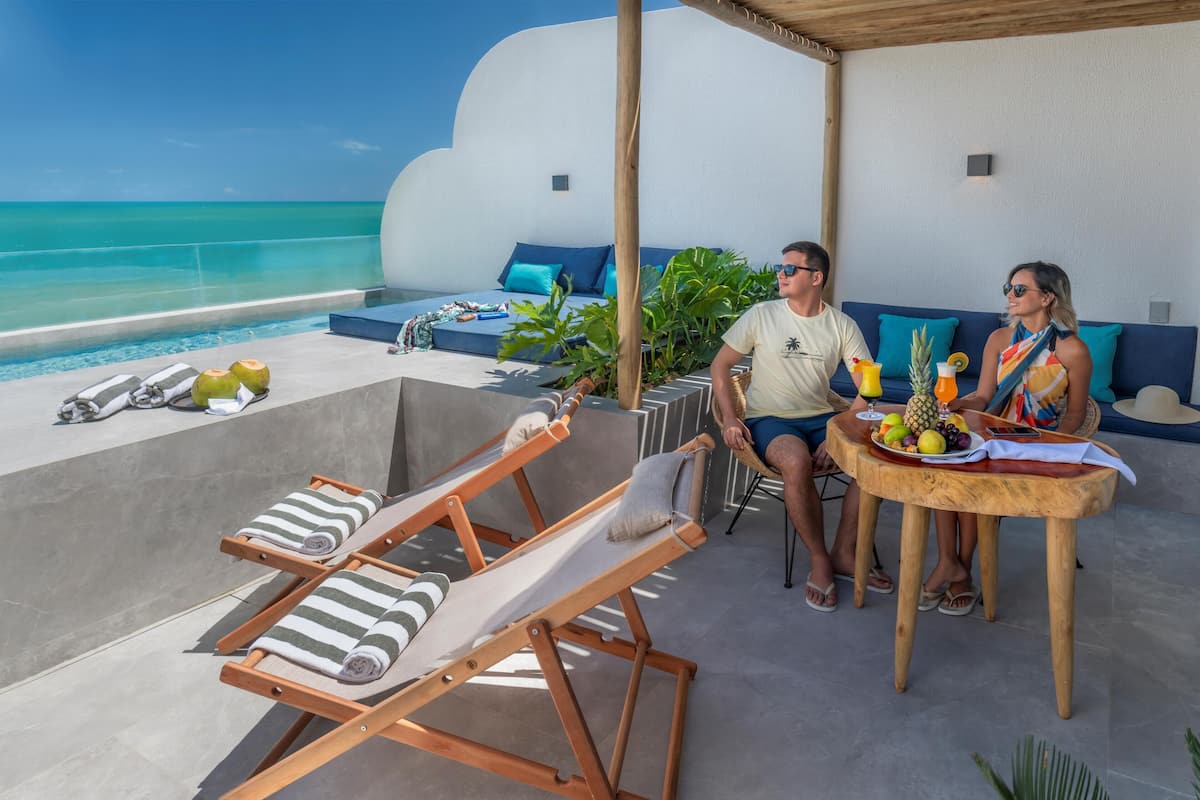 A couple seated at a wooden table with a tropical fruit platter, overlooking a pool with lounge chairs and a view of the ocean.