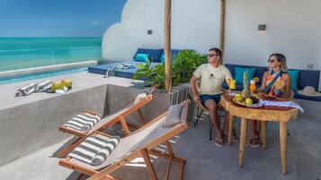 A couple seated at a wooden table with a tropical fruit platter, overlooking a pool with lounge chairs and a view of the ocean.