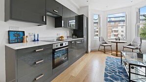 Une cuisine moderne avec des meubles sombres, un plan de travail blanc et un four encastré. On y trouve un coin repas avec une table et des chaises en verre, ainsi que de grandes fenêtres offrant une vue sur la ville.