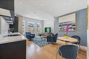 A modern living room with a kitchen area, a dining table, and a large window offering a city view. - Nightingale Suites (London)