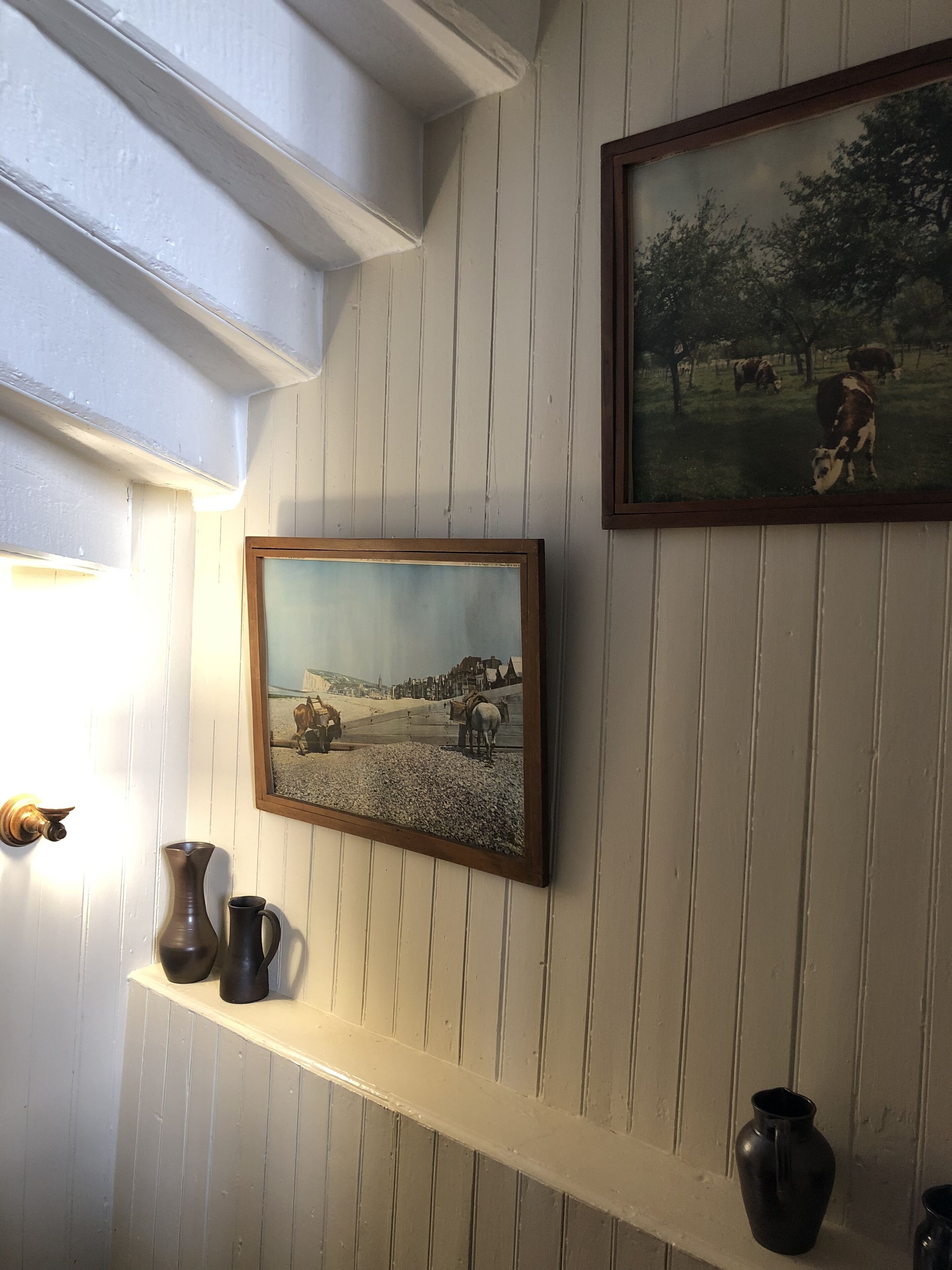 Una habitación con una pared de madera blanca, un cuadro enmarco de una escena campestre y dos jarrones oscuros sobre una estantería.