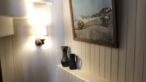 A hallway with a white door, a lit wall sconce, a framed painting, and a shelf with two dark vases. - Maison Ancienne Avec son Jardin Privatif (Le Havre)