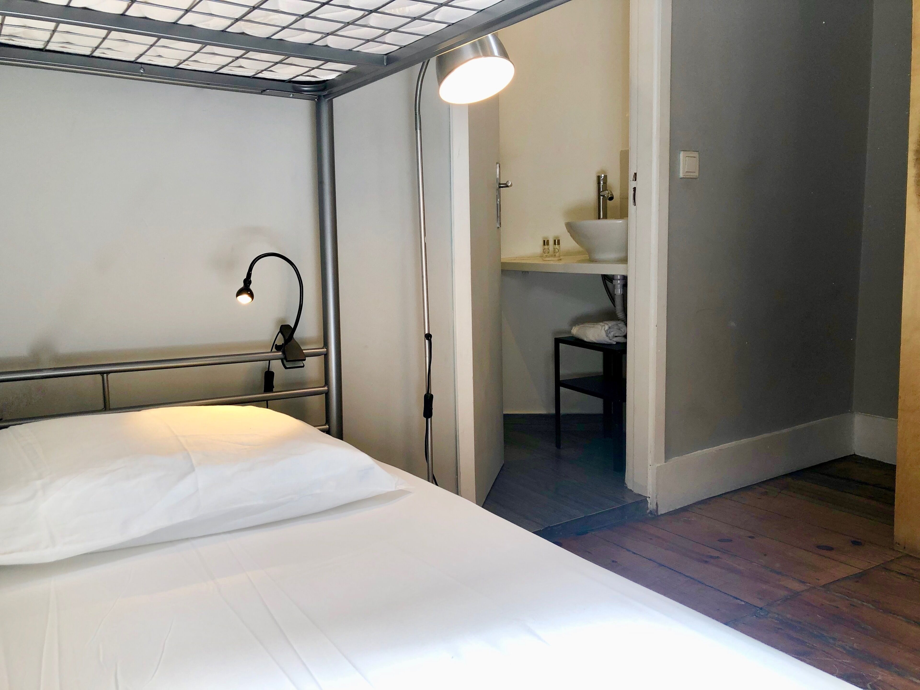 A bedroom with a bed, a desk, a lamp, and a bathroom visible through an open door.