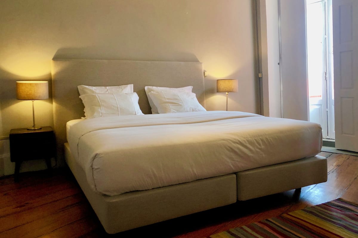 A neatly made bed with white linens and two bedside lamps, a wooden floor, and a door to the right.