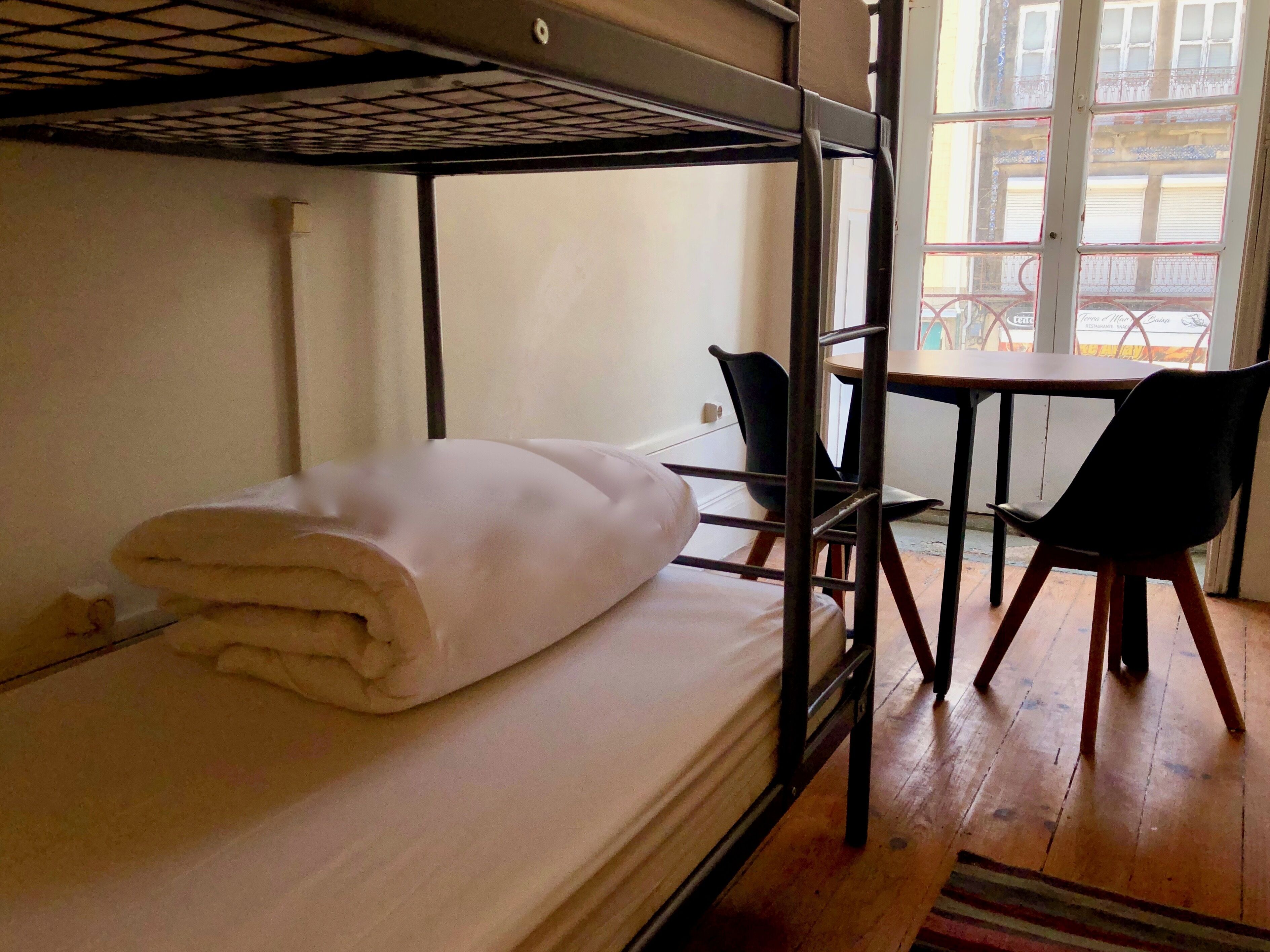 A bunk bed with a rolled white blanket, a small table with two chairs, and a window with a view of buildings.