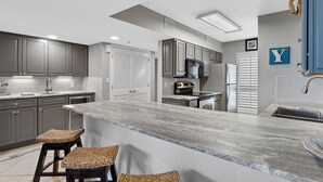A modern kitchen with grey cabinets, a large island with stools, stainless steel appliances, and a blue accent wall. - Mainsail 362 - Gulf Front Condo, Community Pool, Hot Tub, Free Beach Service! (Miramar Beach)