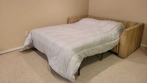 A small, single-bed room with a plain beige carpet, a plain beige wall, and a window with a white frame.