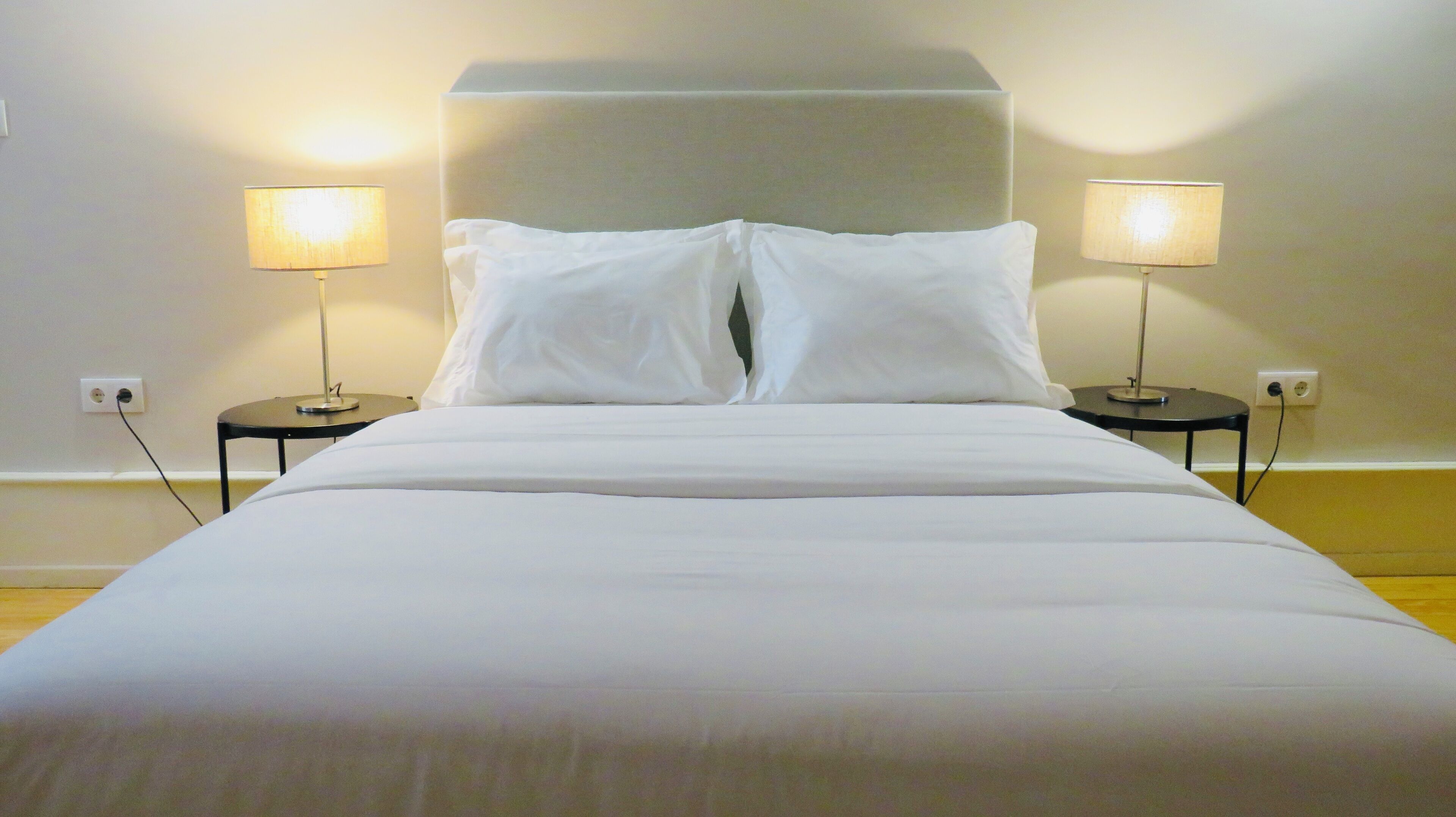 A neatly made bed with white linens, flanked by two bedside tables with lamps, in a room with plain walls and a wooden floor.