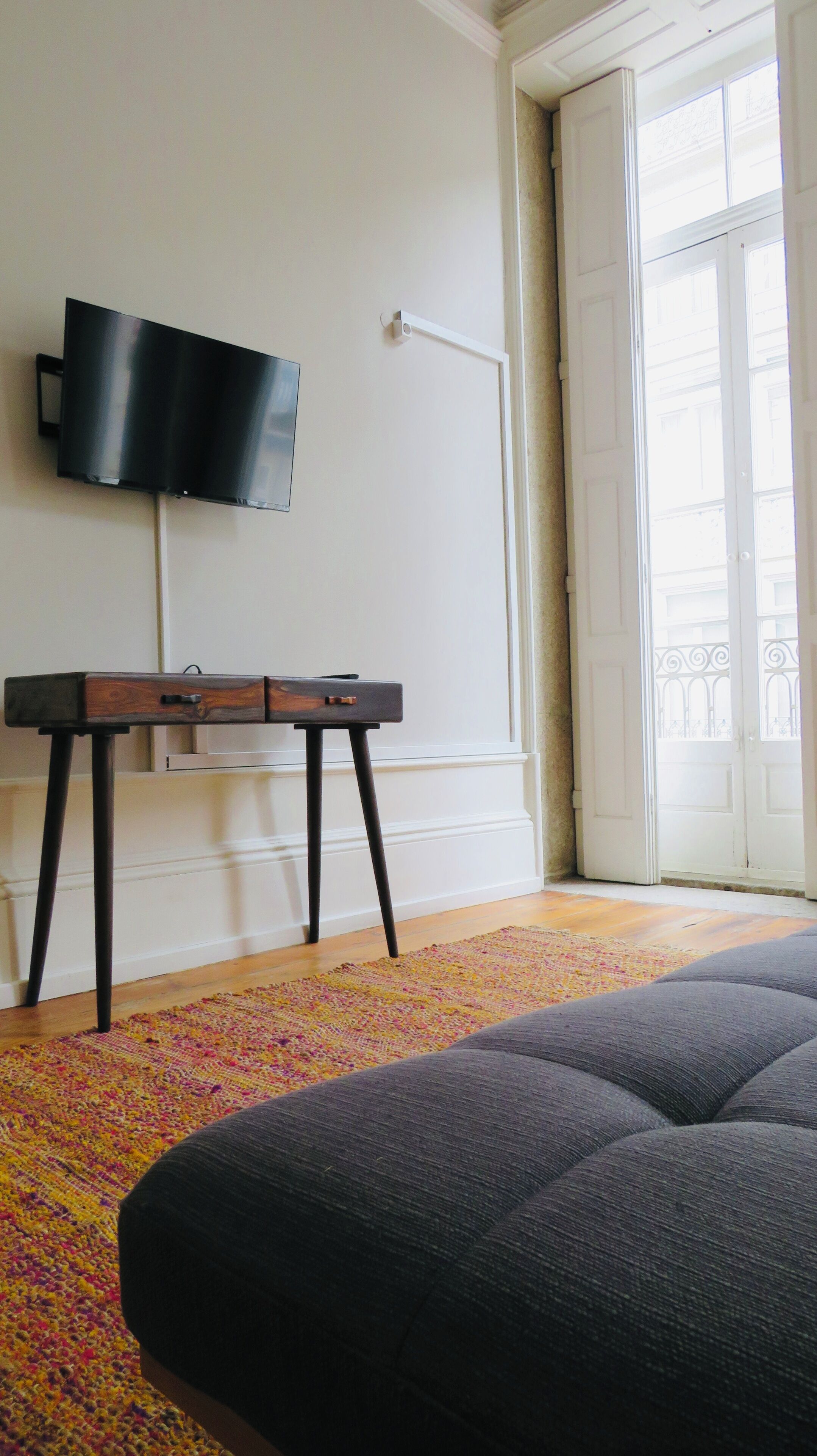 Una stanza con TV a schermo piatto montata a parete, una scrivania in legno con cassetti, un tappeto colorato e un grande divano scuro.
