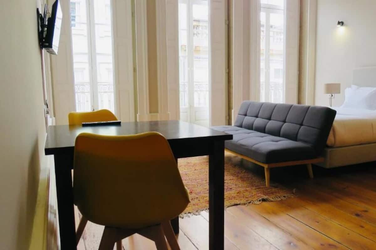 A room with a wooden floor, a grey sofa, a yellow chair, a small table, and a bed with white bedding.