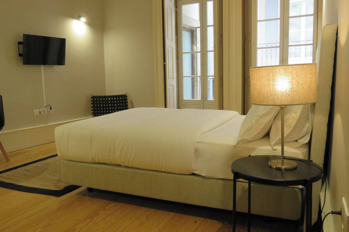 A hotel room with a bed, a desk, a chair, a TV mounted on the wall, and a window with a view of the outside.