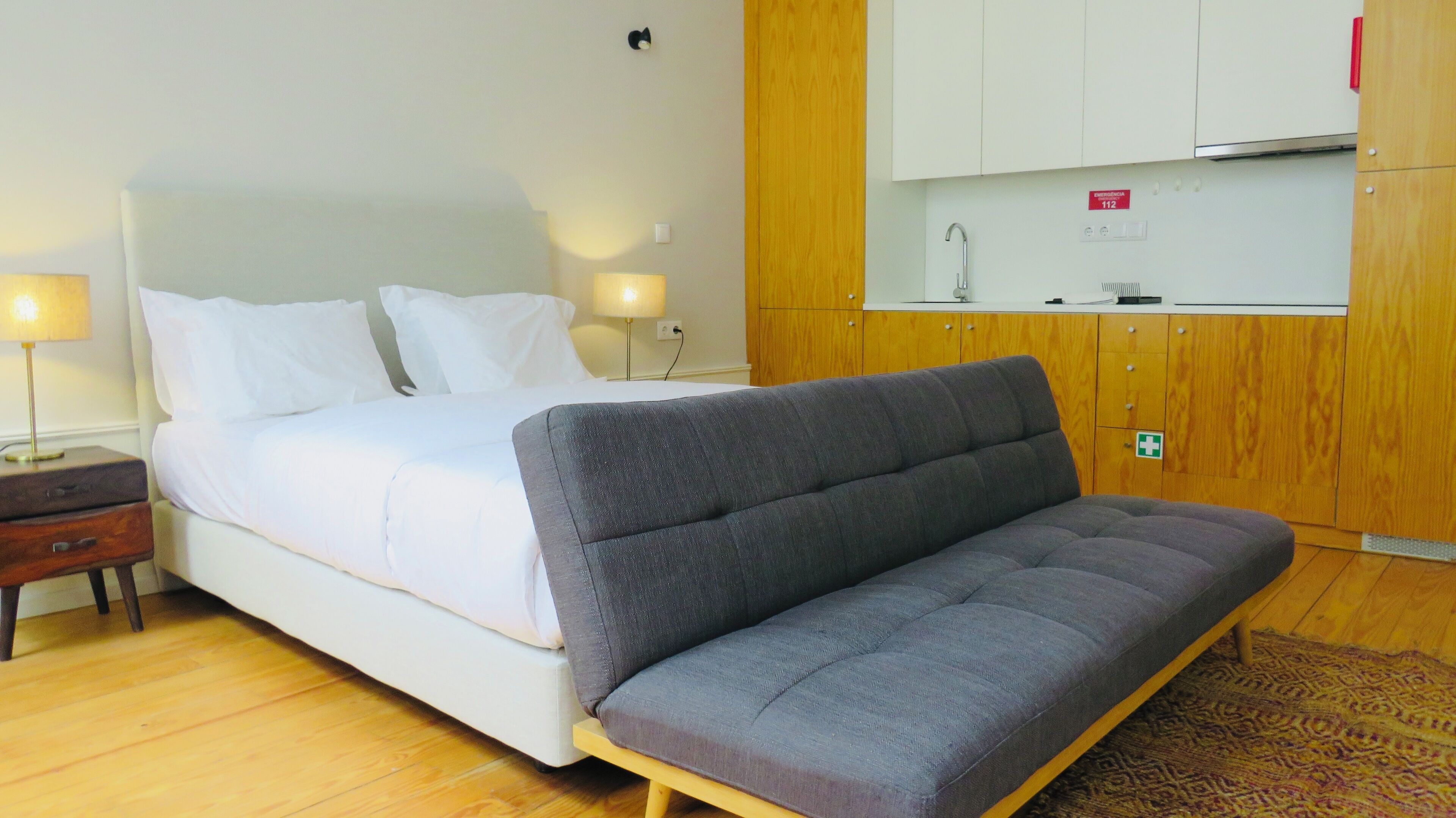 A bedroom with a bed, bedside tables, a sofa, a wooden wardrobe, and a kitchen area with cabinets and a sink.