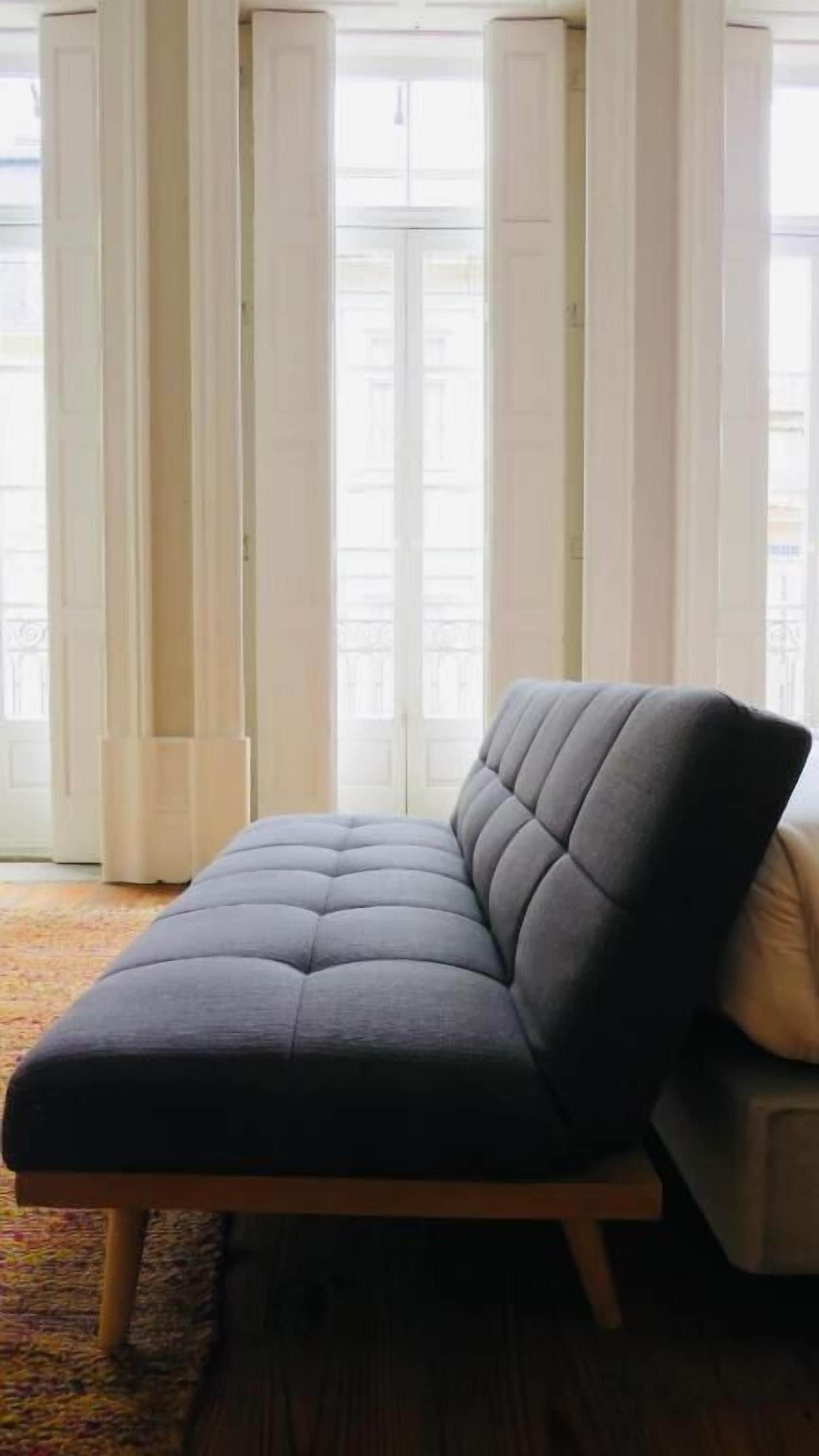 A grey sofa with wooden legs in a room with large windows and white curtains.