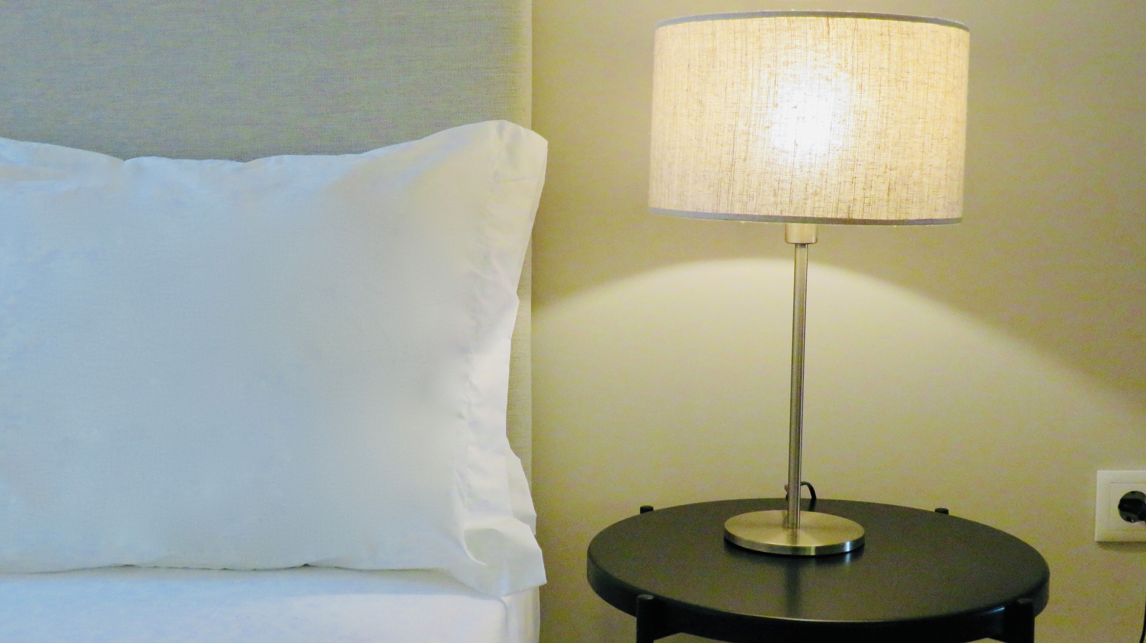 A bed with a white pillow and a round table with a lamp beside it.