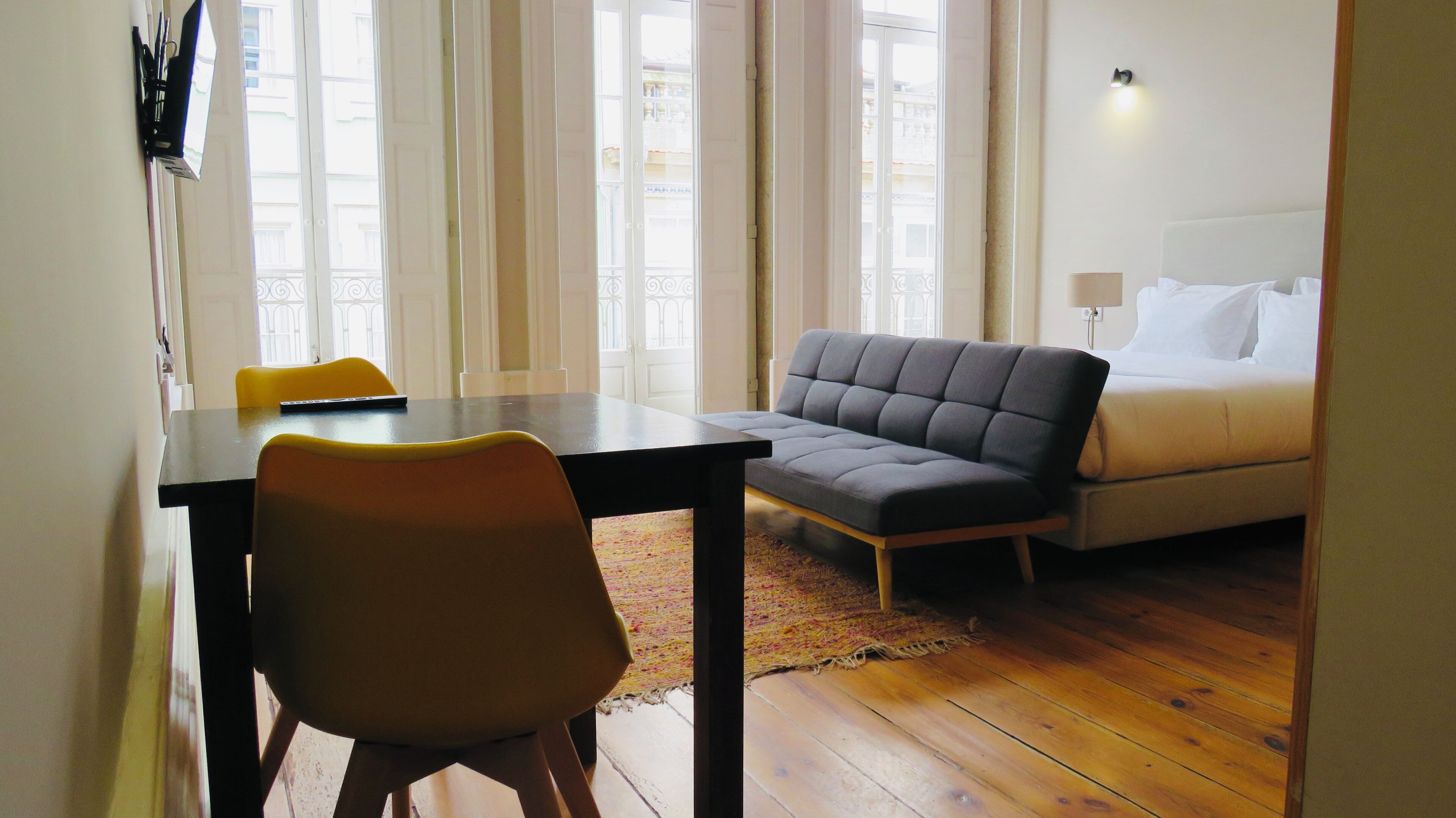 A room with a wooden floor, a grey sofa, a white bed, and a small table with a chair.
