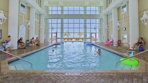 An indoor swimming pool with clear glass walls, a tiled floor, and a few people swimming and relaxing. - Majestic Sun 807B - Stunning Gulf Views, Community Pool, Hot Tub, Exercise Room! (Miramar Beach)