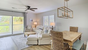 A spacious living room with a ceiling fan, a dining table with chairs, and a view of the outdoors through large windows. - Grayton Beach Villas 101 - Scenic 30A, Steps to the Sand, Walk to Red Bar! (Grayton Beach)