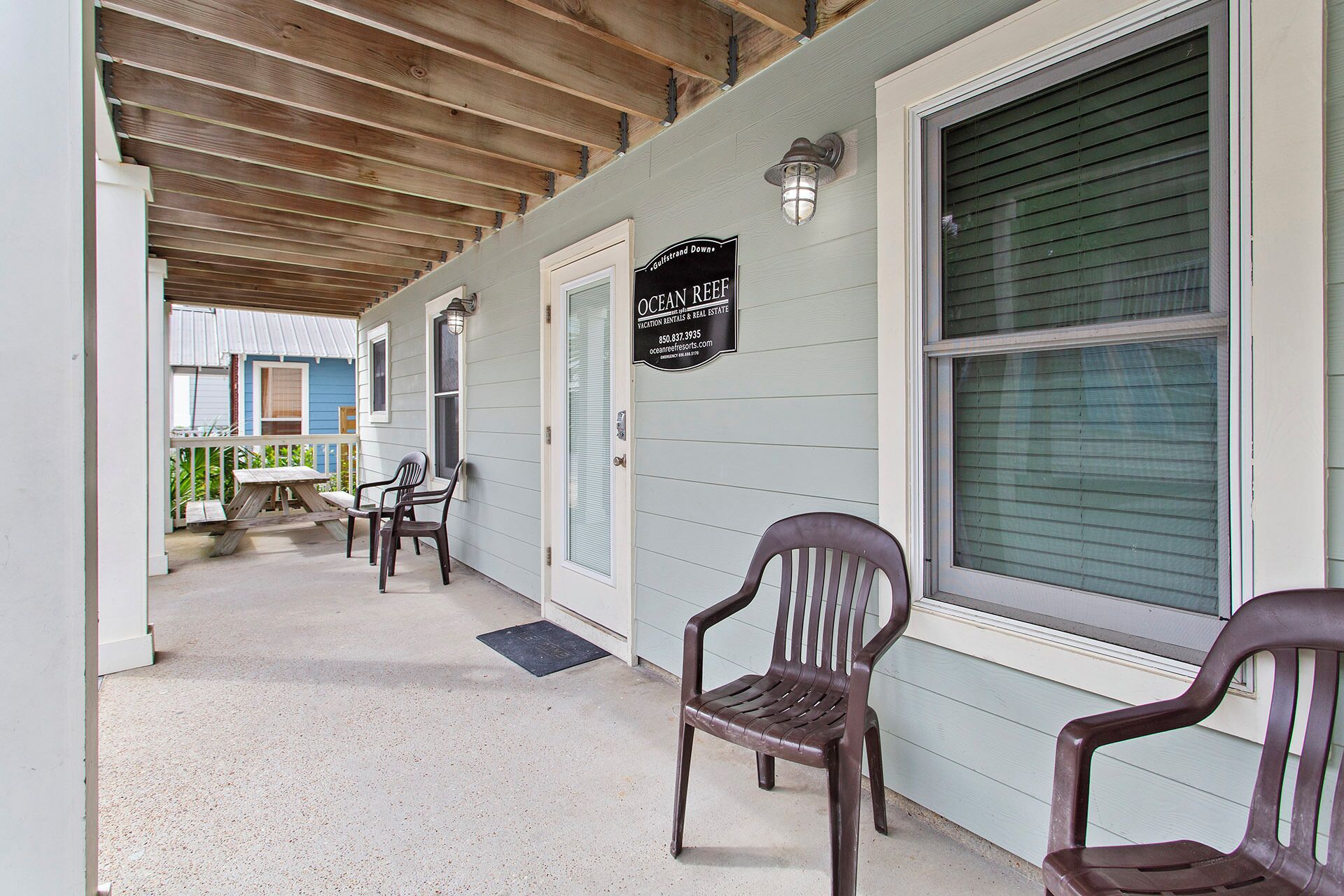 A covered porch with two plastic chairs, a wooden table, and a sign for Ocean Beef Vacation Rentals & Real Estate.