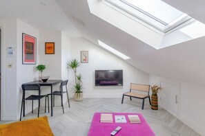Et moderne loftværelse med ovenlys, et fladskærms-tv, et spiseområde med bord og stole, og et siddeområde med sofa og sofabord.