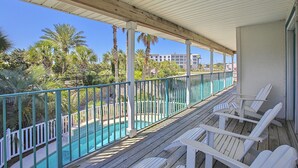 Eine Holzterrasse mit weißen Stühlen mit Blick auf einen Swimmingpool, umgeben von einem türkisblauen Geländer und Palmen.