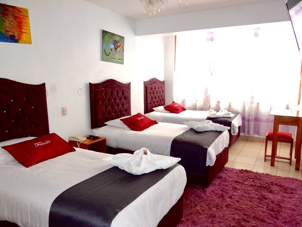 Habitación de hotel con tres camas, cada una con almohada roja y ropa de cama blanca. Hay una mesita de noche de madera con un teléfono y un control remoto. En la pared, un cuadro de colores, y se ve una ventana con cortina morada.