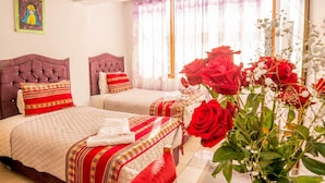 A hotel room with two beds, each with a red and white bedspread, a tufted headboard, and a decorative pillow. There is a large window with sheer curtains and a bouquet of red roses on the windowsill. - Hotel Feimar (Cusco)